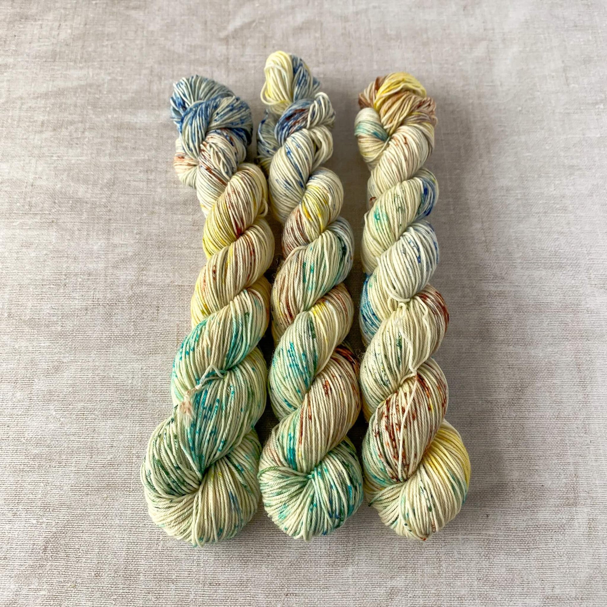 Three skeins of hand-dyed yarn in a light cream color with speckles of green, blue, yellow, and brown, arranged neatly on a textured fabric surface.