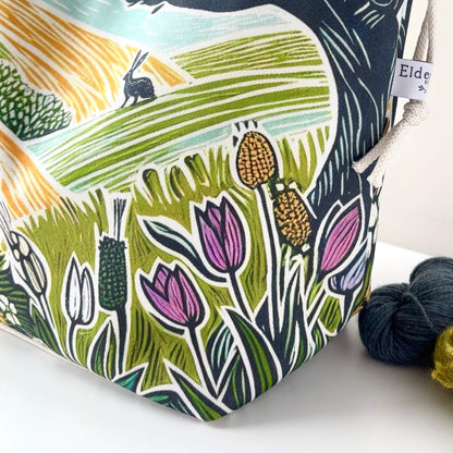 Fabric pouch with nature-themed design featuring flowers and a rabbit, next to yarn balls.