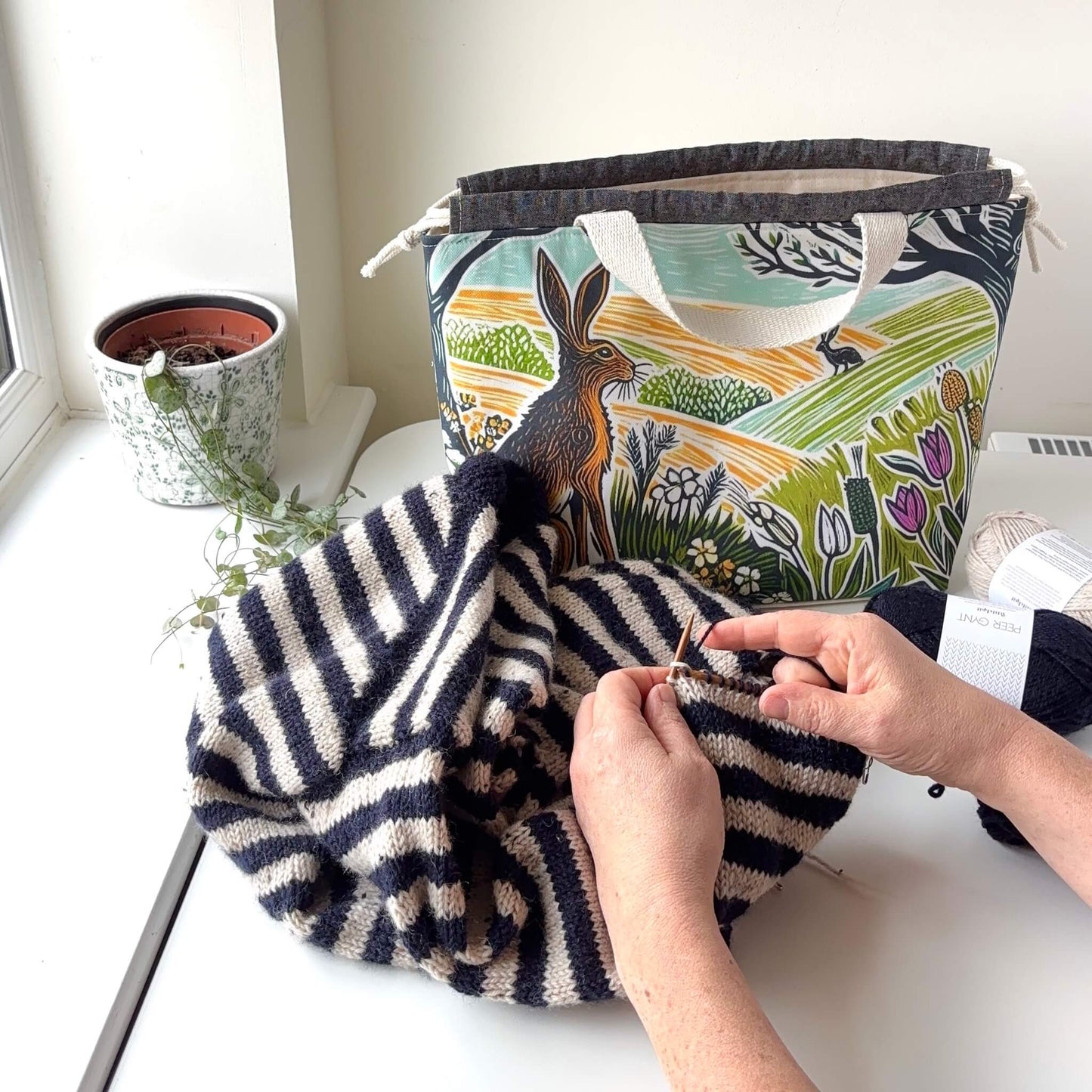 Person knitting with a striped yarn on a white surface, with a patterned bag and plant in the background.
