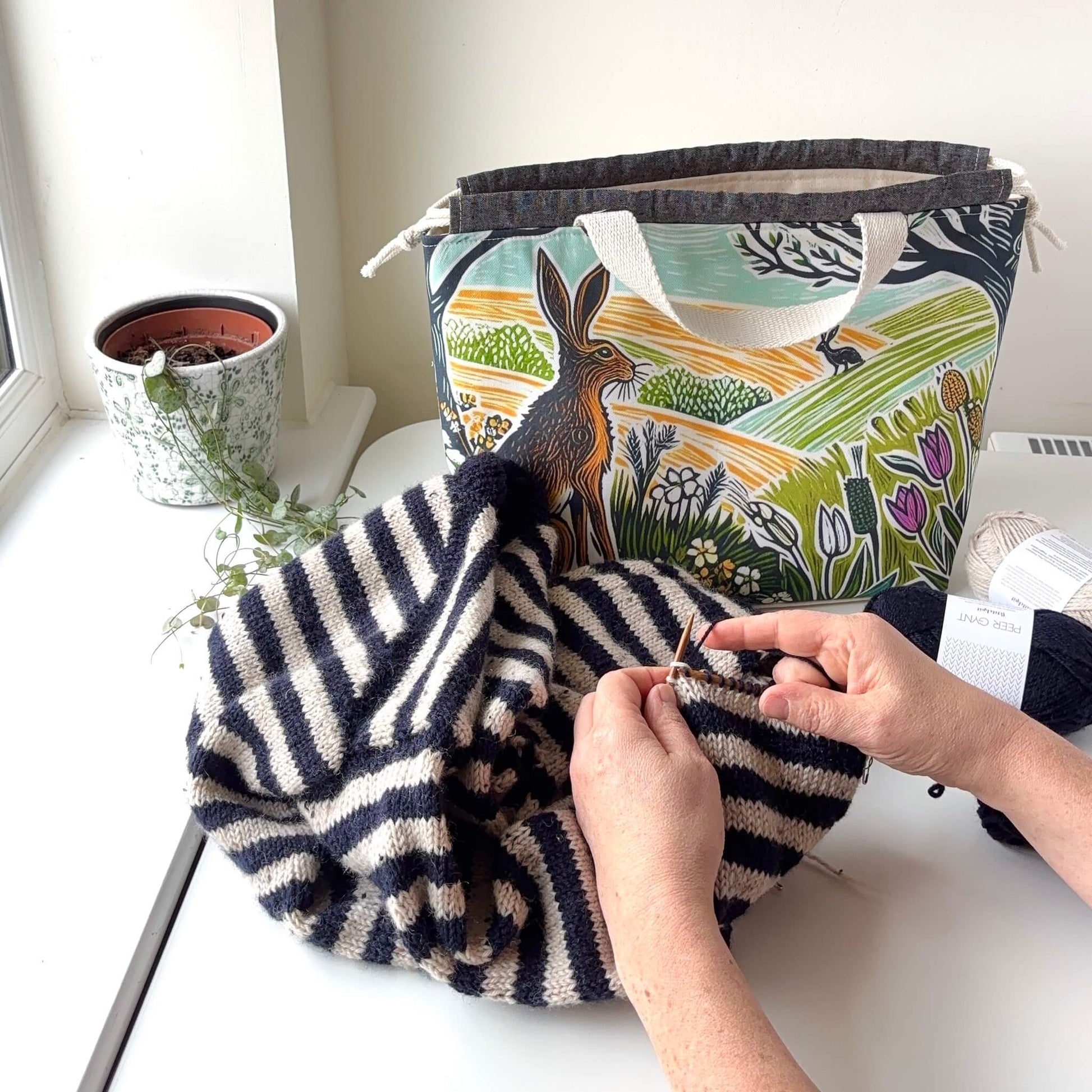Person knitting with a striped yarn on a white surface, with a patterned bag and plant in the background.