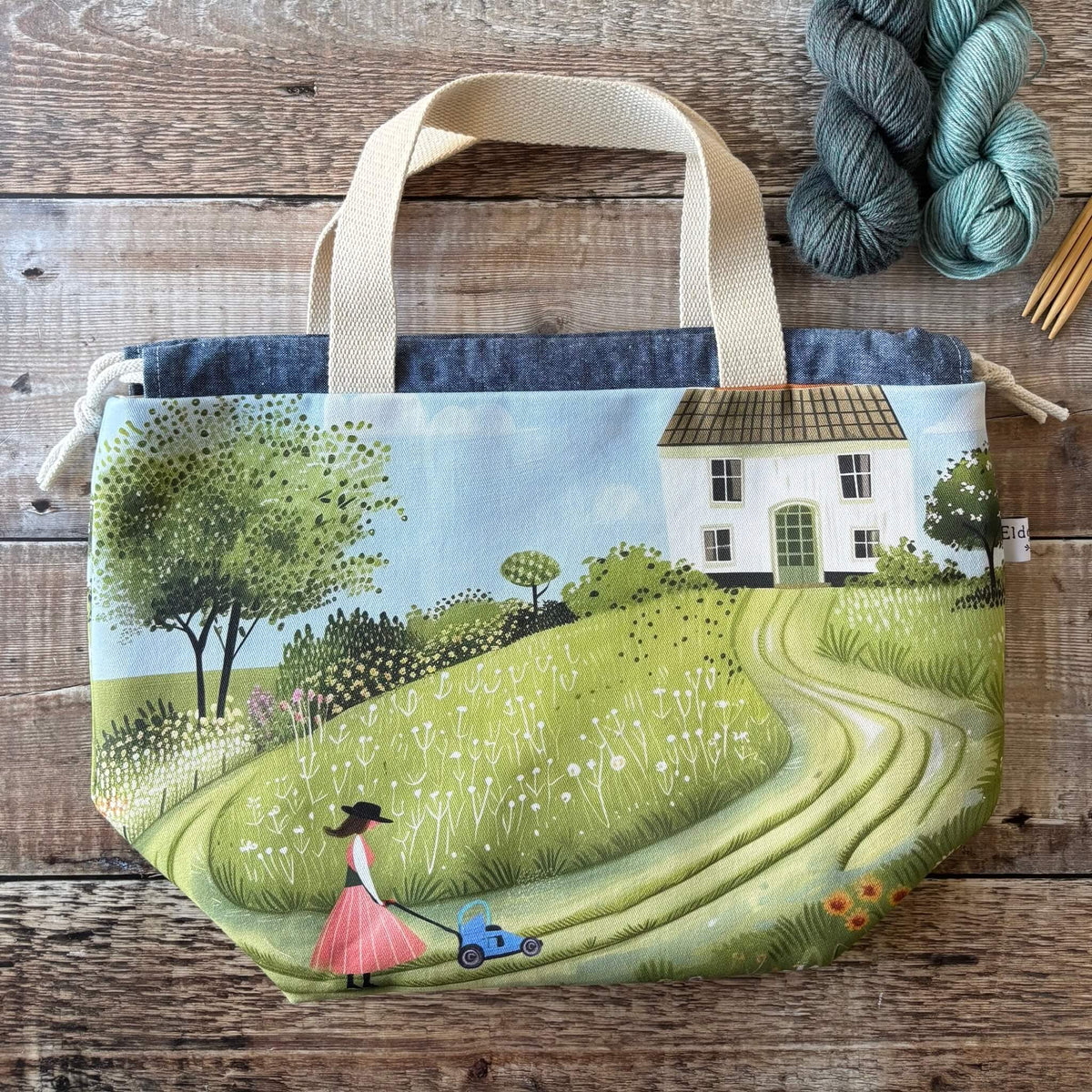 A fabric project bag with a printed illustration of a country scene, featuring a winding green path, a white house, trees, and a woman pushing a blue lawn mower. The bag has cream canvas handles and is displayed on a rustic wooden surface next to skeins of yarn.