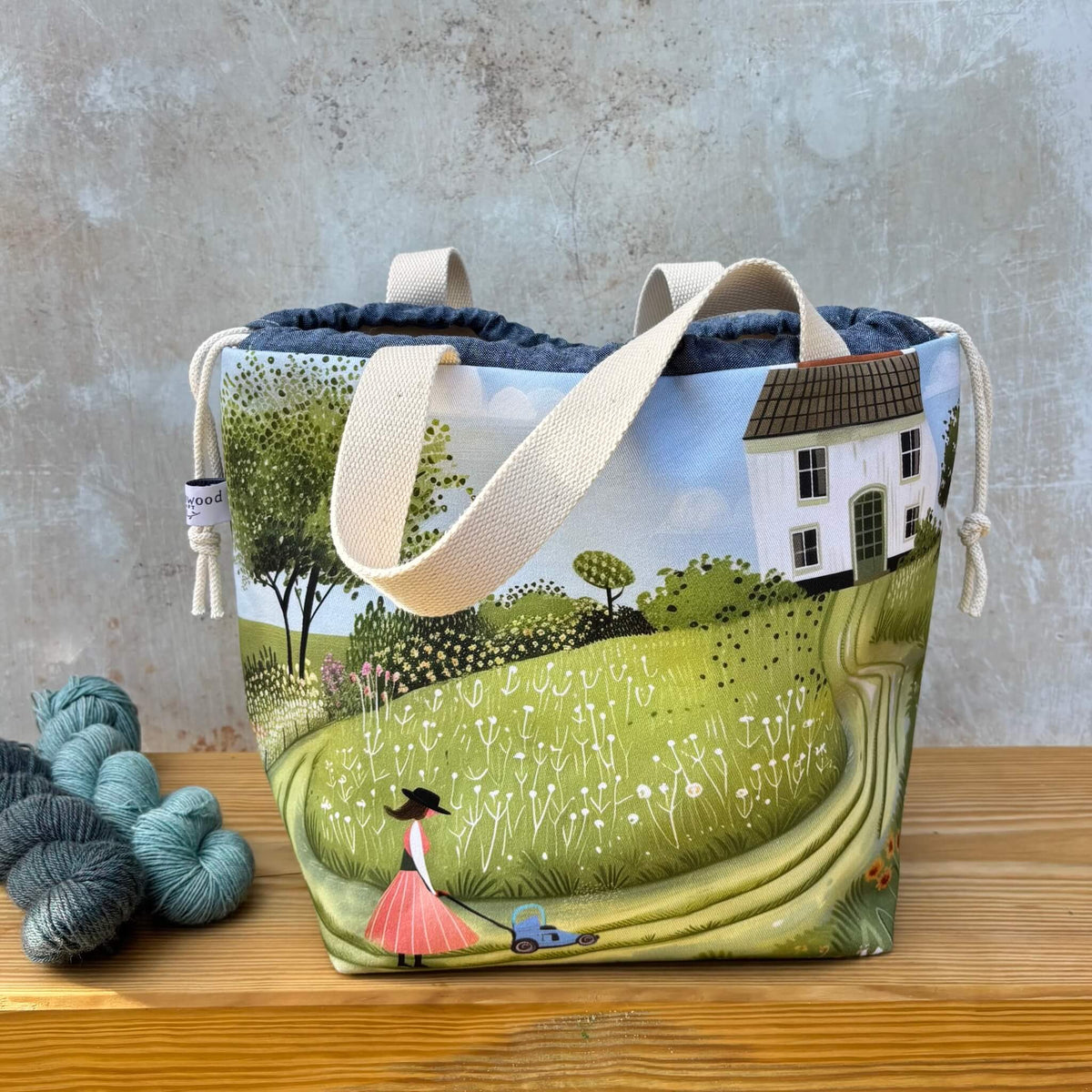 A project bag with a printed landscape design of a woman mowing a lawn in a green field, set against a white house and blue sky. The bag has cream canvas handles and is displayed on a wooden surface next to skeins of yarn against a textured grey wall.
