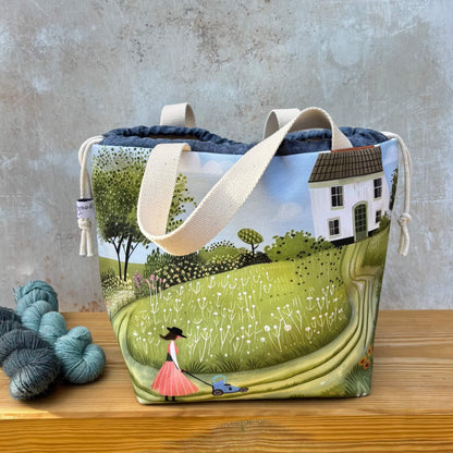 A project bag with a printed landscape design of a woman mowing a lawn in a green field, set against a white house and blue sky. The bag has cream canvas handles and is displayed on a wooden surface next to skeins of yarn against a textured grey wall.