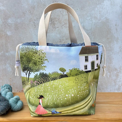 A project bag with cream-coloured handles and a printed illustration of a countryside landscape, featuring a white house, green trees, a grassy field, and a woman pushing a blue lawnmower. The bag is displayed on a wooden surface next to some blue and grey yarn balls against a textured grey wall.