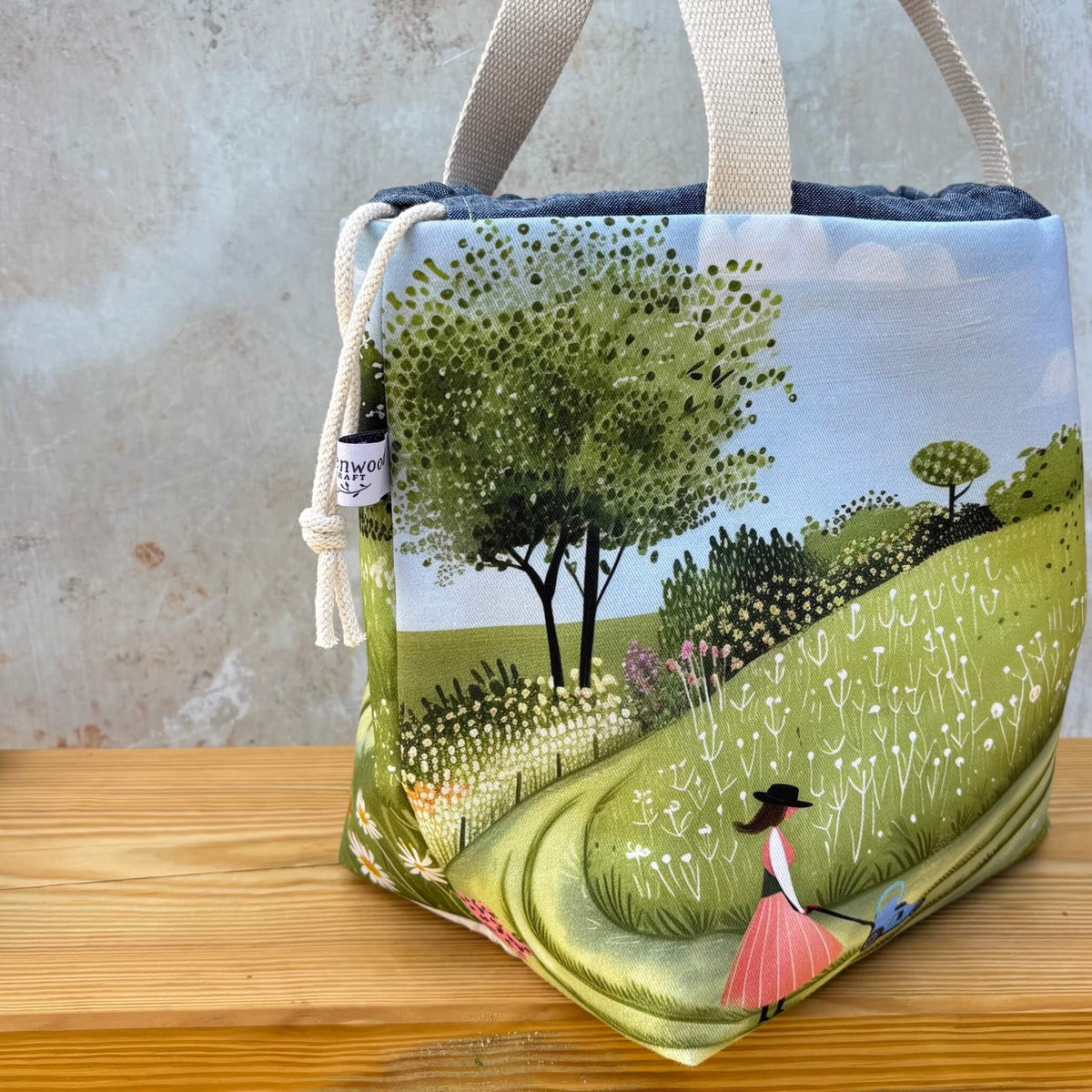 A project bag with a whimsical landscape illustration, featuring a green hill, trees, and a woman in a pink dress. The bag has cream canvas handles and a cream drawstring closure with a small maker's tag that reads Eldenwood Craft.