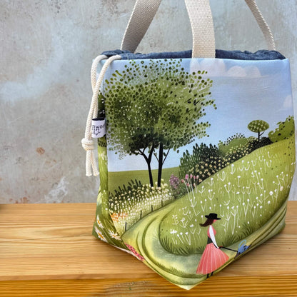 A project bag with a whimsical landscape illustration, featuring a green hill, trees, and a woman in a pink dress. The bag has cream canvas handles and a cream drawstring closure with a small maker's tag that reads Eldenwood Craft.