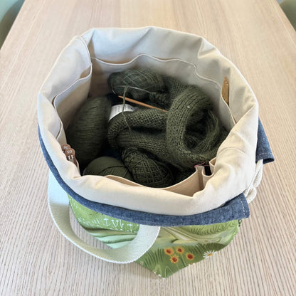 A fabric knitting basket with a cream canvas interior, filled with dark green knitted yarn and knitting needles, set on a light wood table.