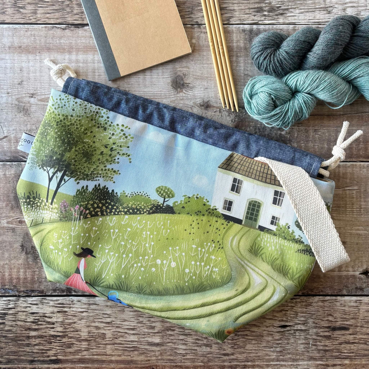 A fabric drawstring pouch with a printed landscape design of a cottage, trees, and a winding path, styled on a rustic wooden surface with knitting needles, yarn, and a notebook.