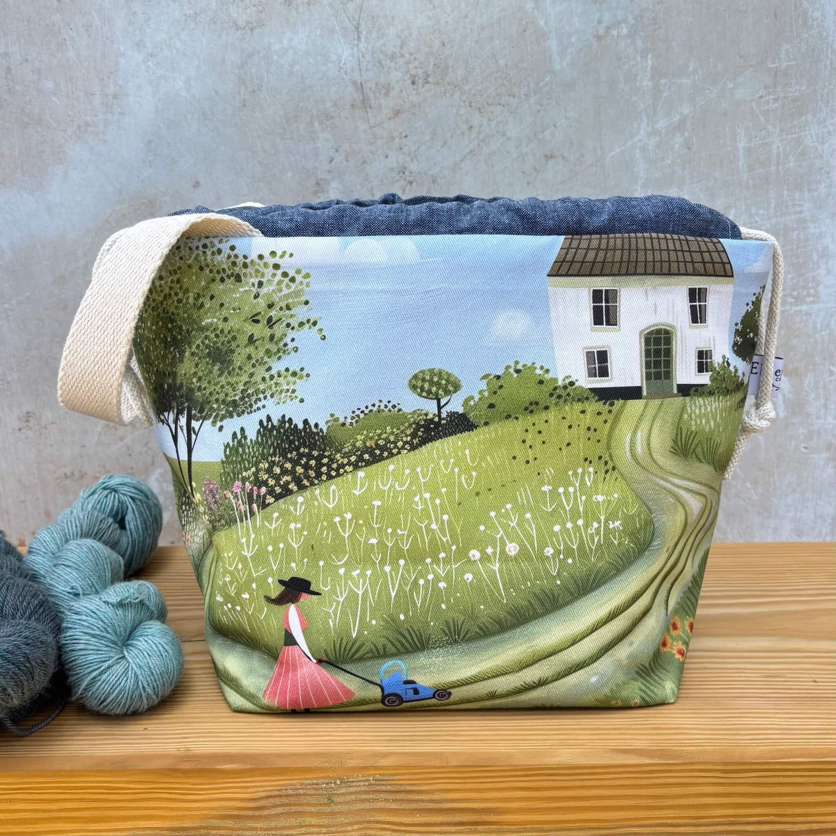 A fabric project bag with a printed countryside scene, featuring a white house, trees, a grassy hill, and a woman mowing her lawn. The bag has a natural beige fabric handle and is displayed on a wooden surface next to skeins of yarn against a textured grey wall.