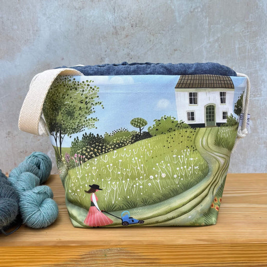 A fabric project bag with a printed countryside scene, featuring a white house, trees, a grassy hill, and a woman mowing her lawn. The bag has a natural beige fabric handle and is displayed on a wooden surface next to skeins of yarn against a textured grey wall.