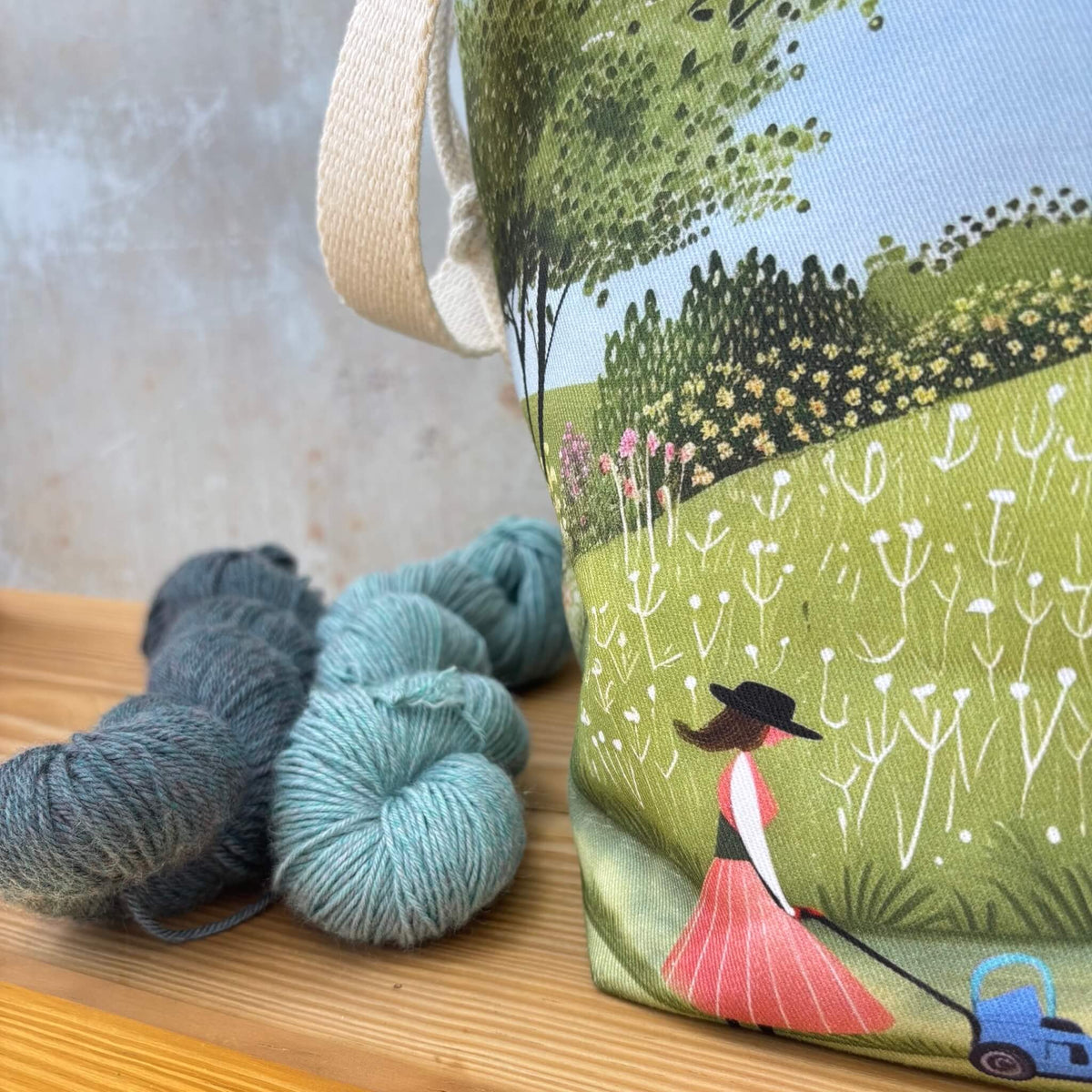 A fabric bag with a whimsical landscape print of a woman mowing a lawn, styled on a wooden surface next to skeins of yarn.