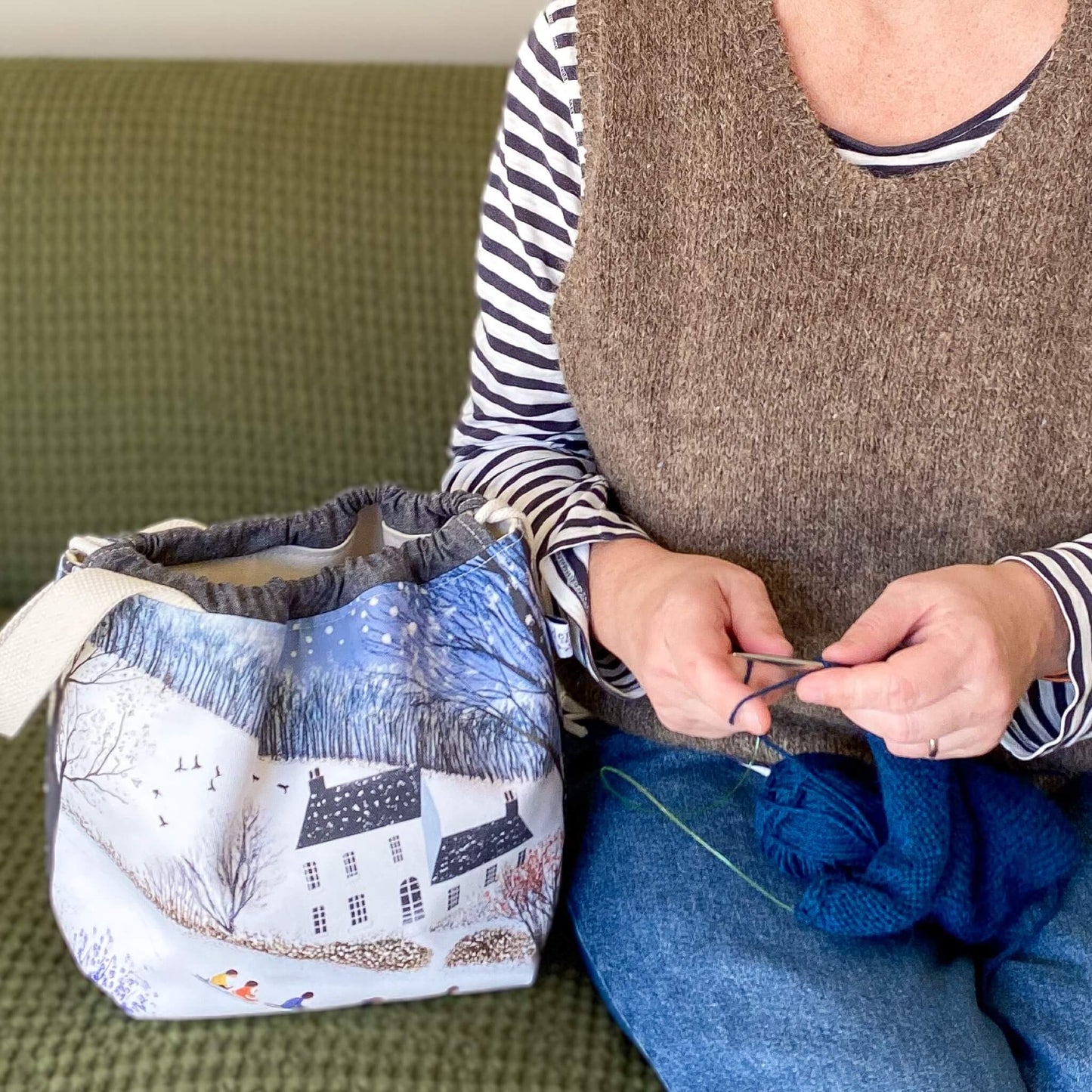 Person knitting with yarn and a winter themed project bag on a green couch