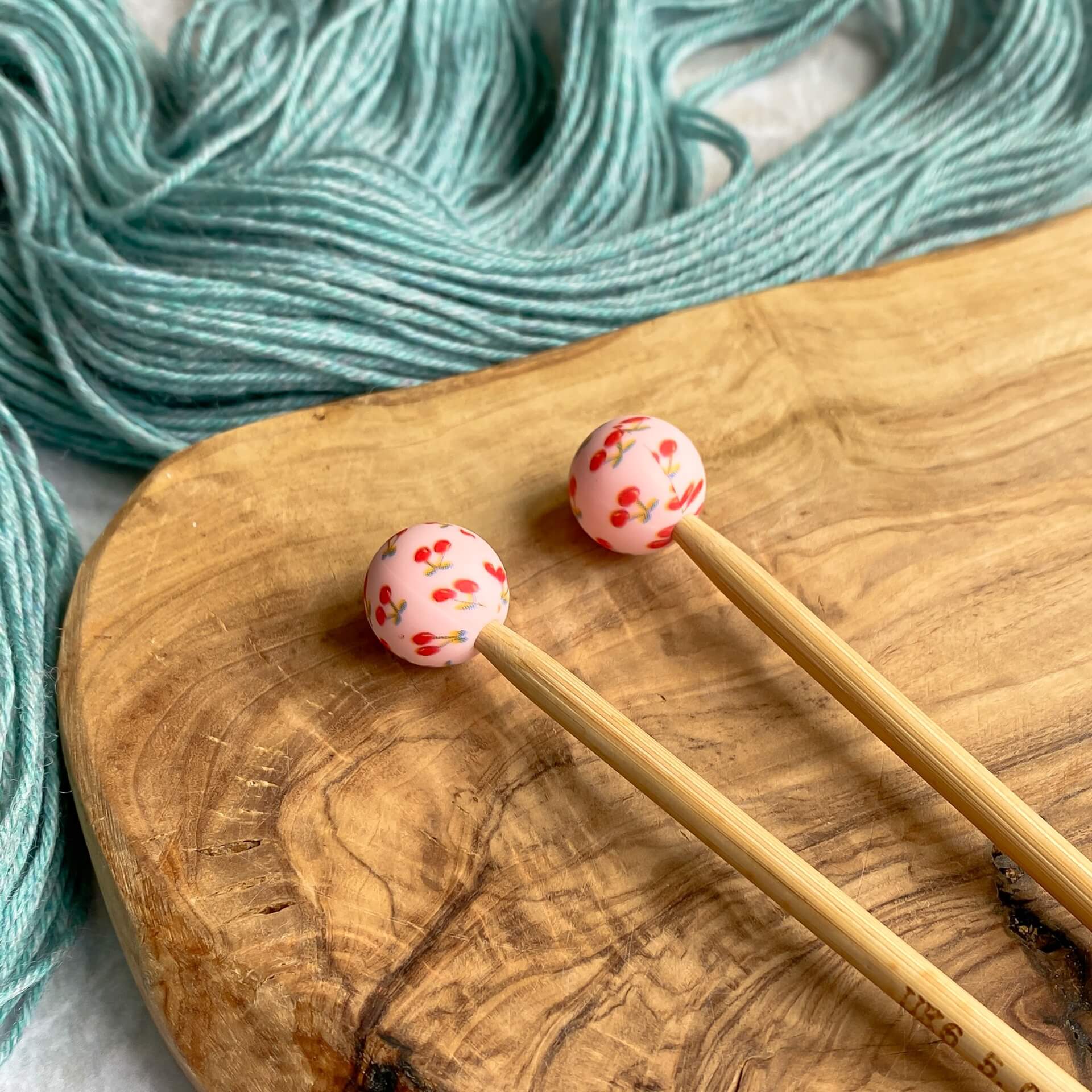 Two silicone balls printed with cherries sit attached to the end of two knitting needles. The needles lie on a wooden surface next to some pale green yarn. 