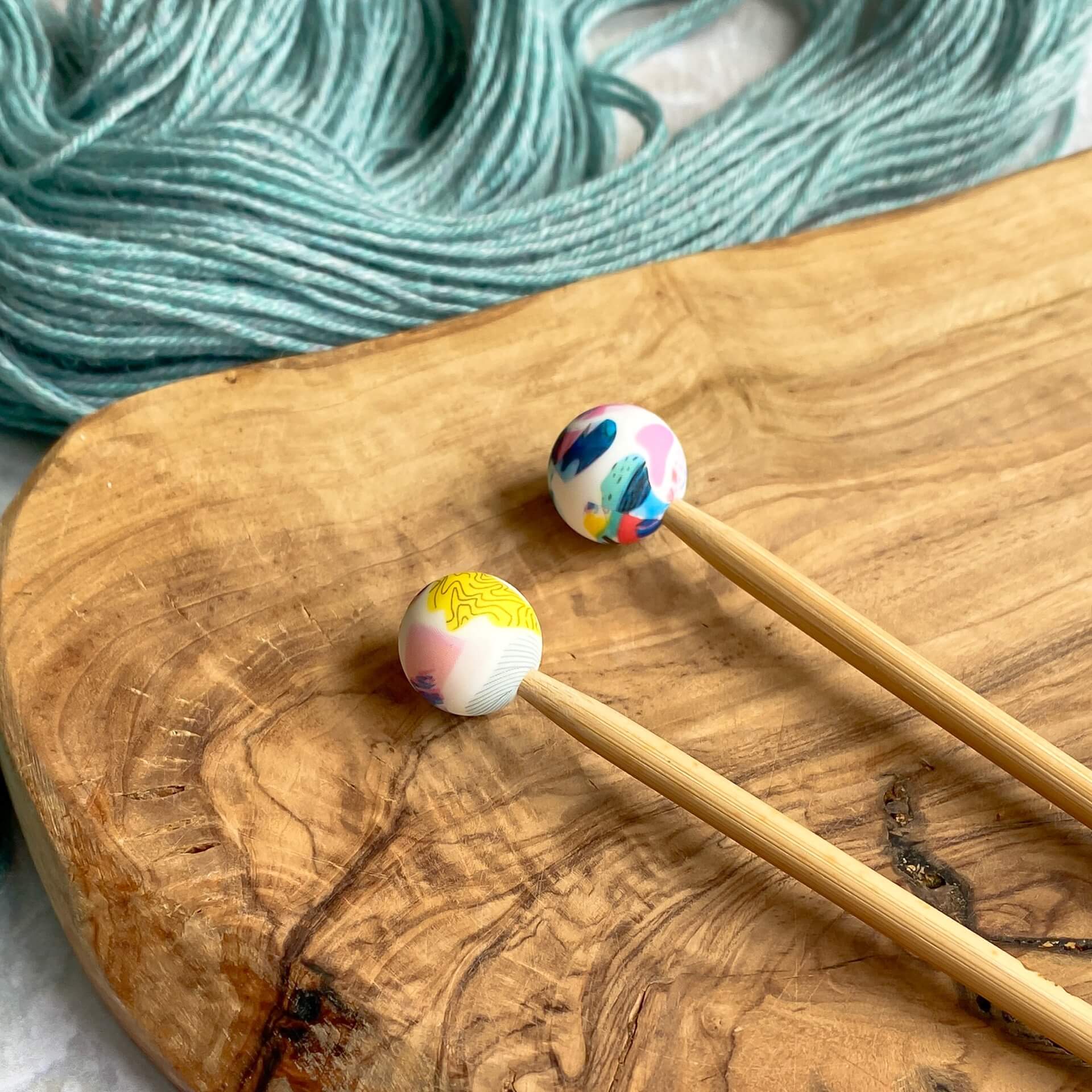 Two silicone balls printed with a bright geometric pattern sit attached to the end of two knitting needles. The needles lie on a wooden surface next to some pale green yarn. 