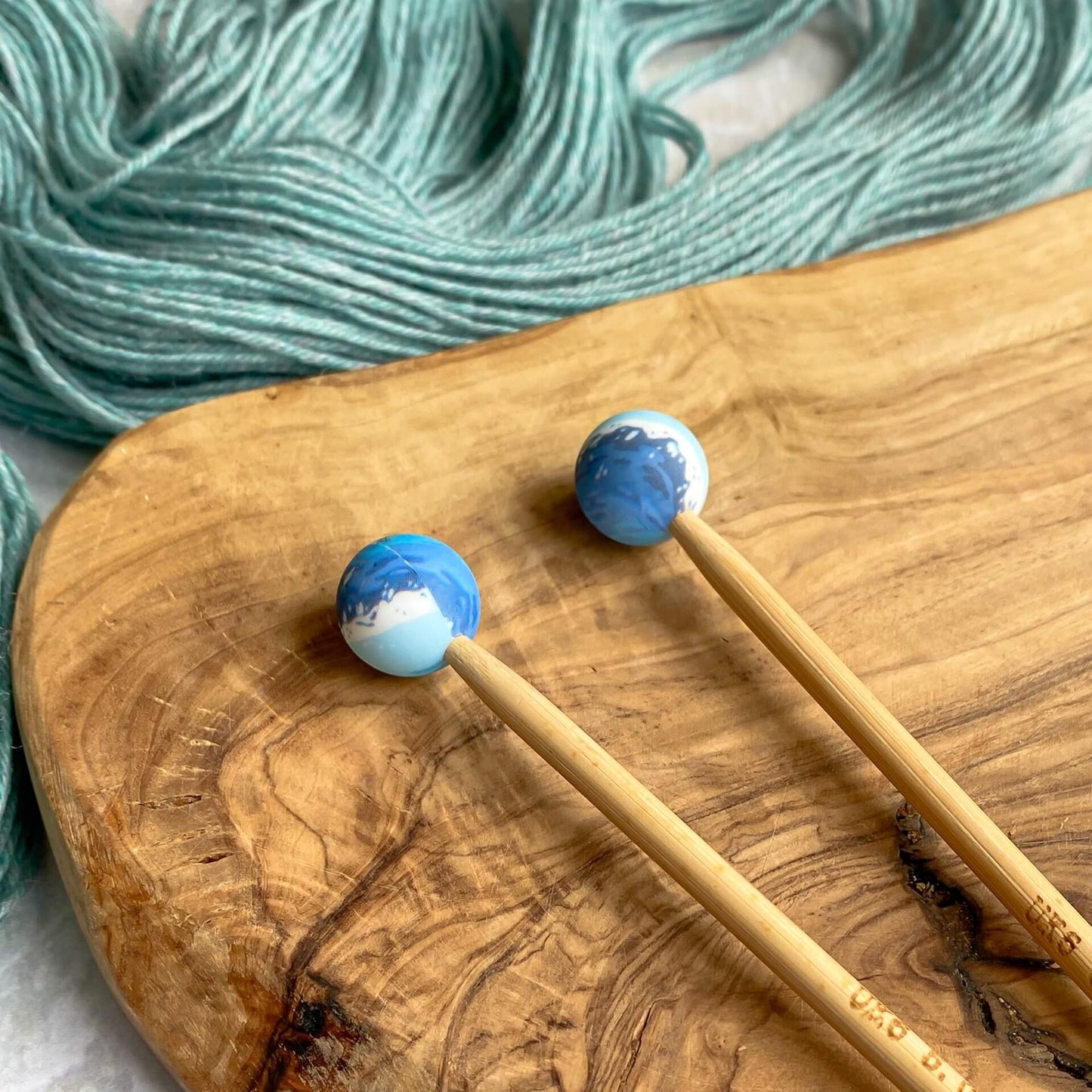 Two silicone balls printed to look like waves sit attached to the end of two knitting needles. The needles lie on a wooden surface next to some pale green yarn. 