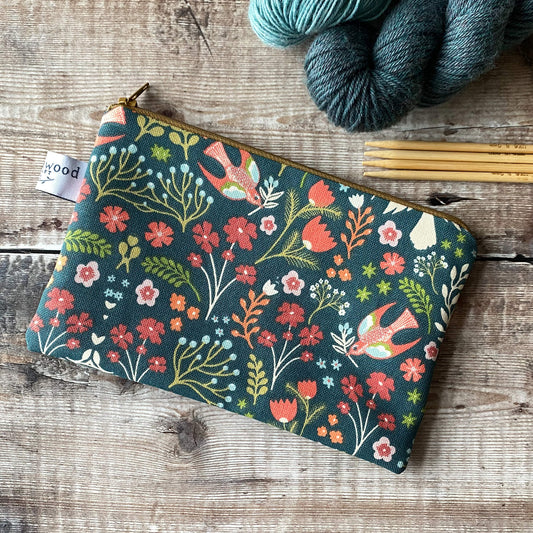 
ChatGPT
The image features a small zippered pouch with a floral pattern, showcasing vibrant flowers and birds in shades of red, orange, and teal on a dark green background. The pouch is accompanied by several skeins of yarn and knitting needles, which are placed on a wooden surface. The pouch appears to be designed for storing knitting notions, and its zipper and fabric design are visible in detail.