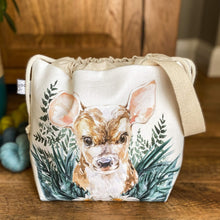 Load image into Gallery viewer, A project bag for knitting is seen on a wooden floor. The bag is a drawstring bag and on the fabric is a baby deer with big ears sitting in amongst greenery