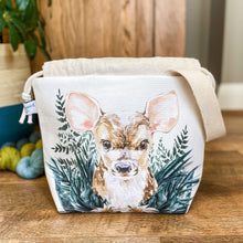 Load image into Gallery viewer, A project bag for knitting is seen on a wooden floor. The bag is a drawstring bag and on the fabric is a baby deer with big ears sitting in amongst greenery. Behind the bag can be seen a large green houseplant and some yarn on the floor. The top of the drawstring is open. 