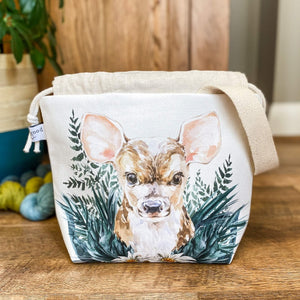 A project bag for knitting is seen on a wooden floor. The bag is a drawstring bag and on the fabric is a baby deer with big ears sitting in amongst greenery. Behind the bag can be seen a large green houseplant and some yarn on the floor. The top of the drawstring is open. 