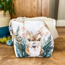 Load image into Gallery viewer, A project bag for knitting is seen on a wooden floor. The bag is a drawstring bag and on the fabric is a baby deer with big ears sitting in amongst greenery with some yarn and a house plant in the background.