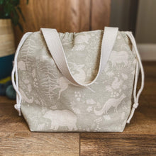 Load image into Gallery viewer, We are looking at a large knitting project bag, pulled closed by its drawstring. It&#39;s handles are dangling in front of the bag. The bag sits on a wooden floor and next to a house plant and some hanks of yarn. 
