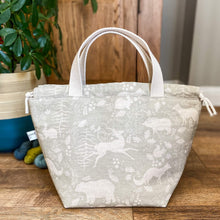 Load image into Gallery viewer, A large knitting project bag is sitting in front of some blue and green yarn and a house plant that is sitting in a beige and teal coloured plant pot. The bag is made from a sage green coloured fabric that is printed with some woodland imagery. 