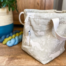 Load image into Gallery viewer, A sage green project bag for craft sits sideways on, next to three skeins of yarn - two blue and one green. There is a houseplant in the background that is sitting in a beige and teal blue plant pot. 