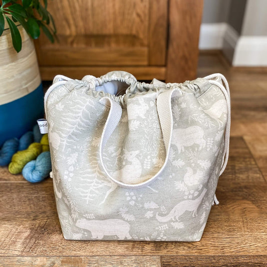 A large drawstring bag is sitting on a wooden floor. The bag has two cream coloured handles and is made with a sage green woodland print. It is designed for use as a knitting or crochet project bag. Next to the bag are some skeins of yarn and a house plant. 