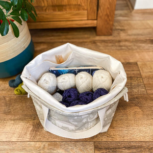 We are looking from the top into a crafter's project bag. It has several skeins of yarn inside as well as a notions pouch and some wooden knitting needles. Also next to the bag are some other skeins of yarn. The bag sits next to a large plant pot. 