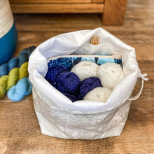Load image into Gallery viewer, A knitting project bag, full of yarn and a notions pouch and wooden knitting needles, sits open on a floor. Next to the bag are three skeins of yarn and a plant pot can be seen in the corner of the image. 