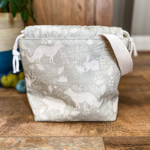 Load image into Gallery viewer, A small knitting project bag is sitting on a wooden floor. The fabric that the bag is made from is a gentle sage green colour and shows woodland animals and flora. 