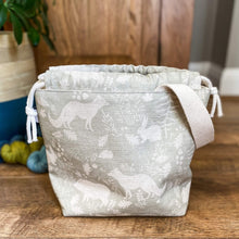 Load image into Gallery viewer, A small knitting project bag is sitting on a wooden floor. The fabric that the bag is made from is a gentle sage green colour and shows woodland animals and flora. Behind the bag are some skeins of yarn and a large house plant.