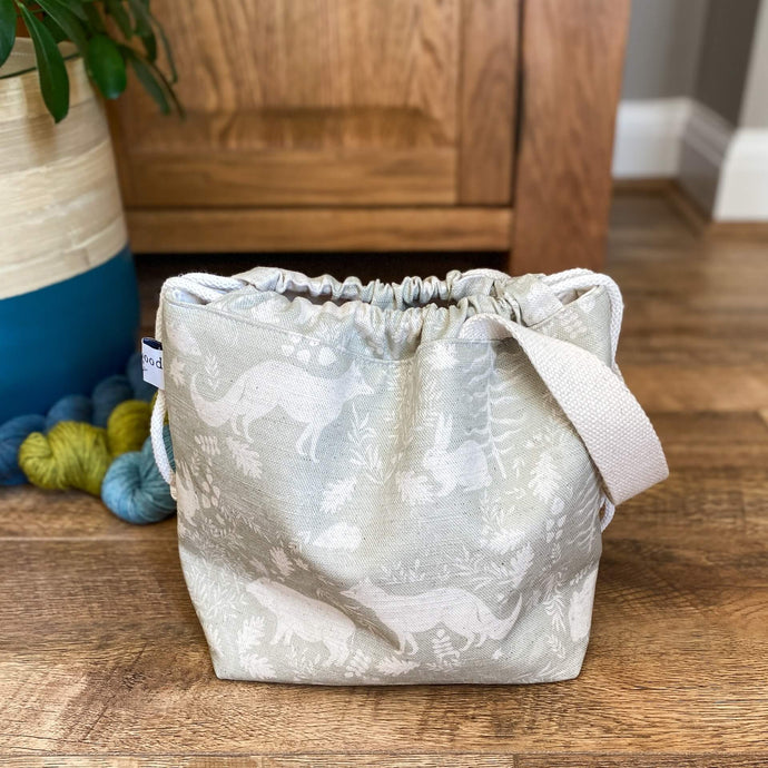 A small knitting project bag with a closed drawstring top is sitting on a wooden floor. The fabric that the bag is made from is a gentle sage green colour and shows woodland animals and flora. 
