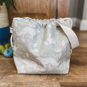 A small knitting project bag is sitting on a wooden floor. The fabric that the bag is made from is a gentle sage green colour and shows woodland animals and flora. 