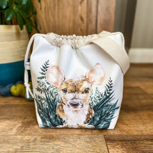 Load image into Gallery viewer, A sweet little baby deer adorns the front of a knitting project bag, which is sitting on a dark wood floor. The deer is nestled in amongst dark green leaves. 