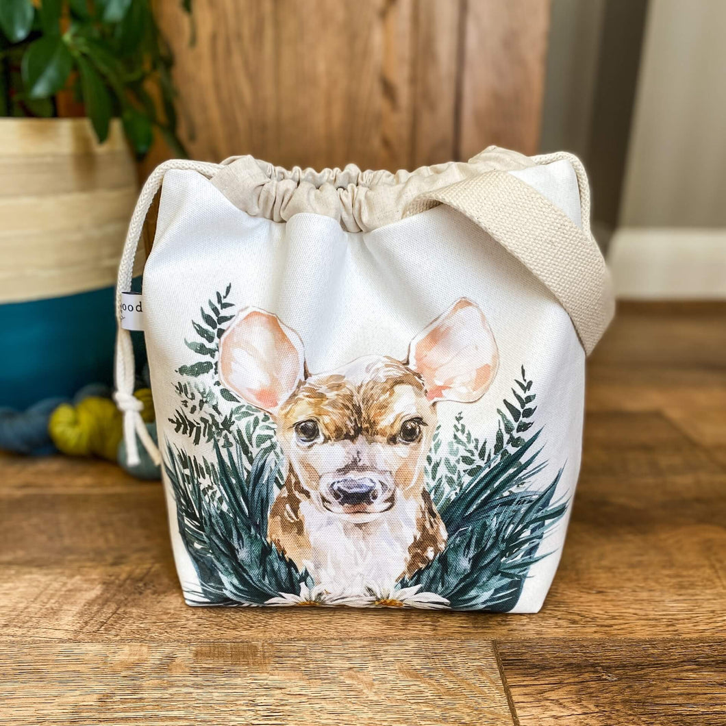 A sweet little baby deer adorns the front of a knitting project bag, which is sitting on a dark wood floor. The deer is nestled in amongst dark green leaves. 