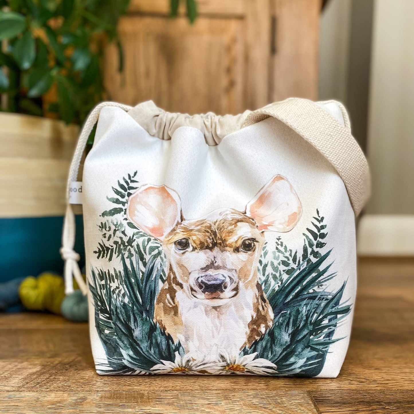A close up of a project bag with a drawstring top. We are looking right into the eyes of a cute little baby deer that is hiding amongst woodland greenery. Behind the bag are some skeins of yarn on the floor and a large houseplant. 