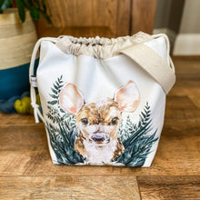 Load image into Gallery viewer, A sweet little baby deer adorns the front of a knitting project bag, which is sitting on a dark wood floor. The deer is nestled in amongst woodland greenery.