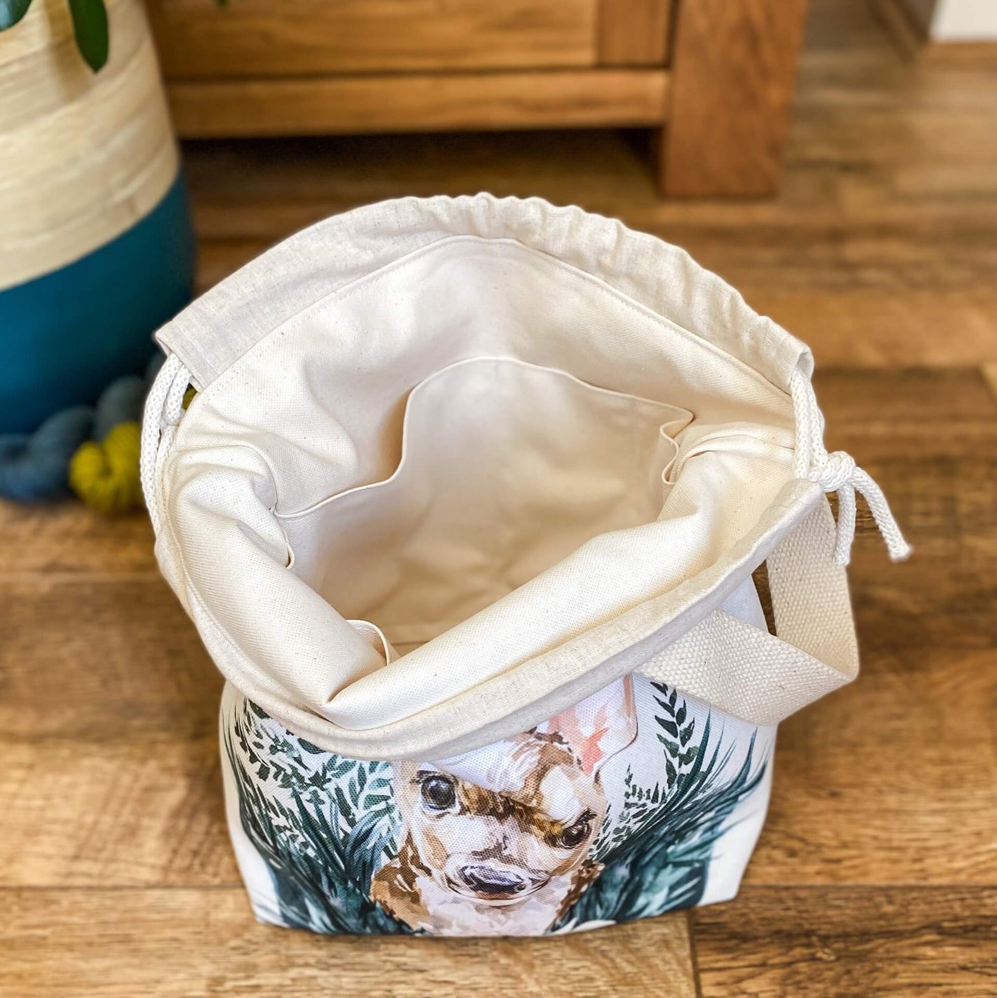 A project bag for knitting is sitting on a wooden floor. It is wide open to show the inside lining and pockets. Behind the bag can be seen two skeins of yarn and a houseplant. 
