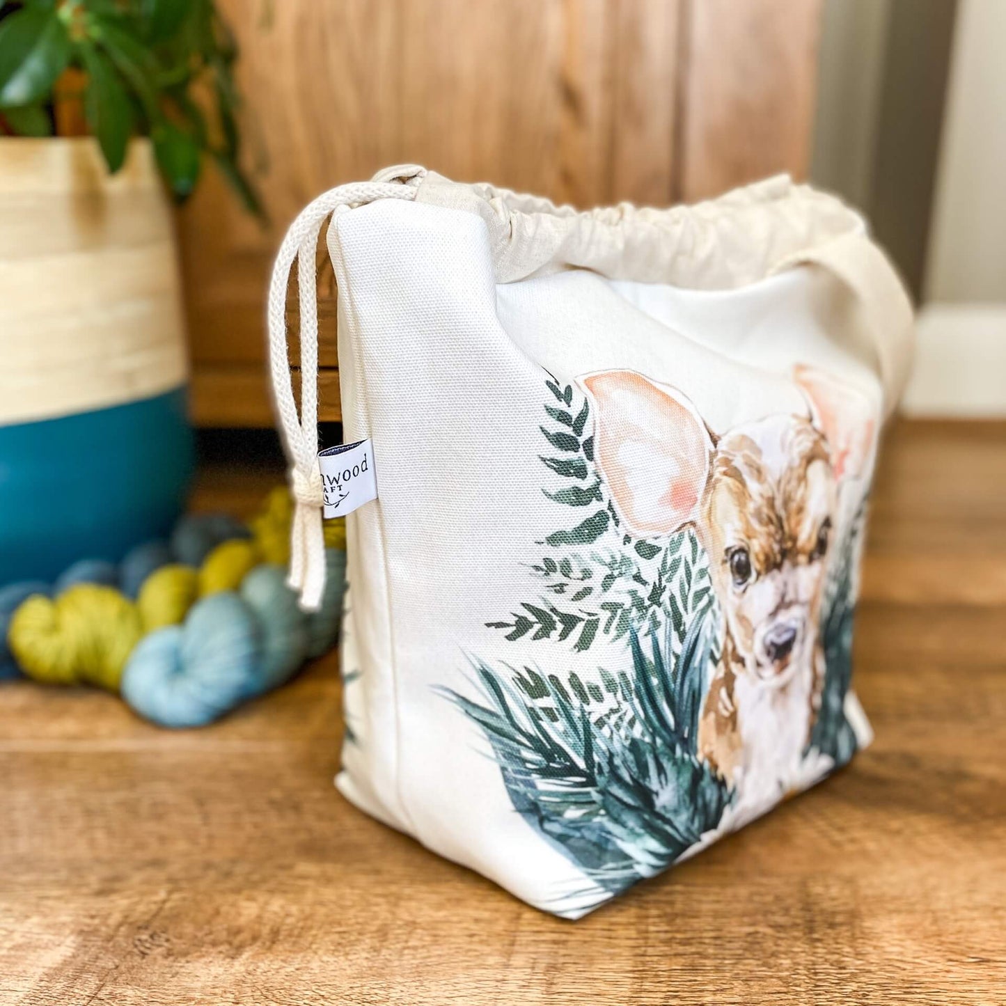 A project bag for knitting is sitting side on on a wooden floor. Next to the bag, which has a cute baby deer on the front, are three skeins of yarn and a large house plant. 