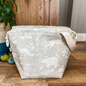 A knitting project bag sits opened on the floor. It's sage green in colour and has woodland animals on it. There are some skeins of yarn behind the bag as well as a house plant. 