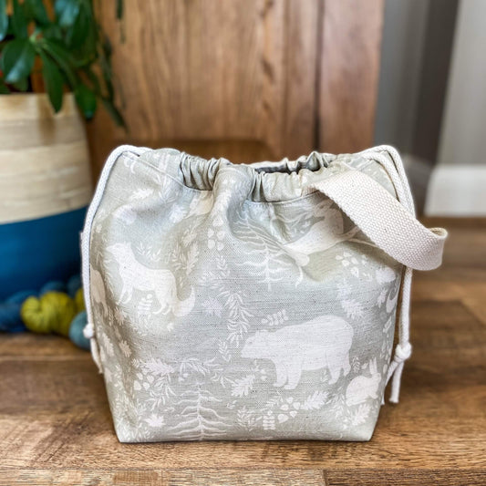 A drawstring project bag is found on a wooden floor. It is pulled closed. The bag is made from a sage green fabric that has woodland animals and flora printed on it. Behind the bag are three skeins of yarn and a house plant. 