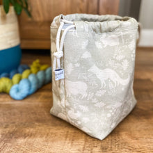 Load image into Gallery viewer, We can see a side on view of a knitting project bag. The bag is a sage green colour and has woodland animals and flora printed on it. Next to the bag are some skeins of yarn and a house plant just in the corner of the view. 