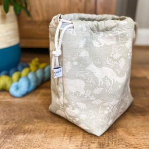 We can see a side on view of a knitting project bag. The bag is a sage green colour and has woodland animals and flora printed on it. Next to the bag are some skeins of yarn and a house plant just in the corner of the view. 