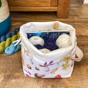 A wide open project bag sits on the floor. We can see inside at the blue and white balls of yarn. There's a notions pouch tucked inside and some wooden knitting needles too. Next to the bag can be seen three skeins of yarn, two blue and one green. 