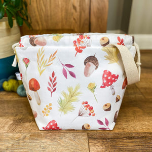 An autumnal knitting project bag is standing open although we can't see inside. The fabric is printed with colourful autumnal imagery such as toadstools and acorns. 