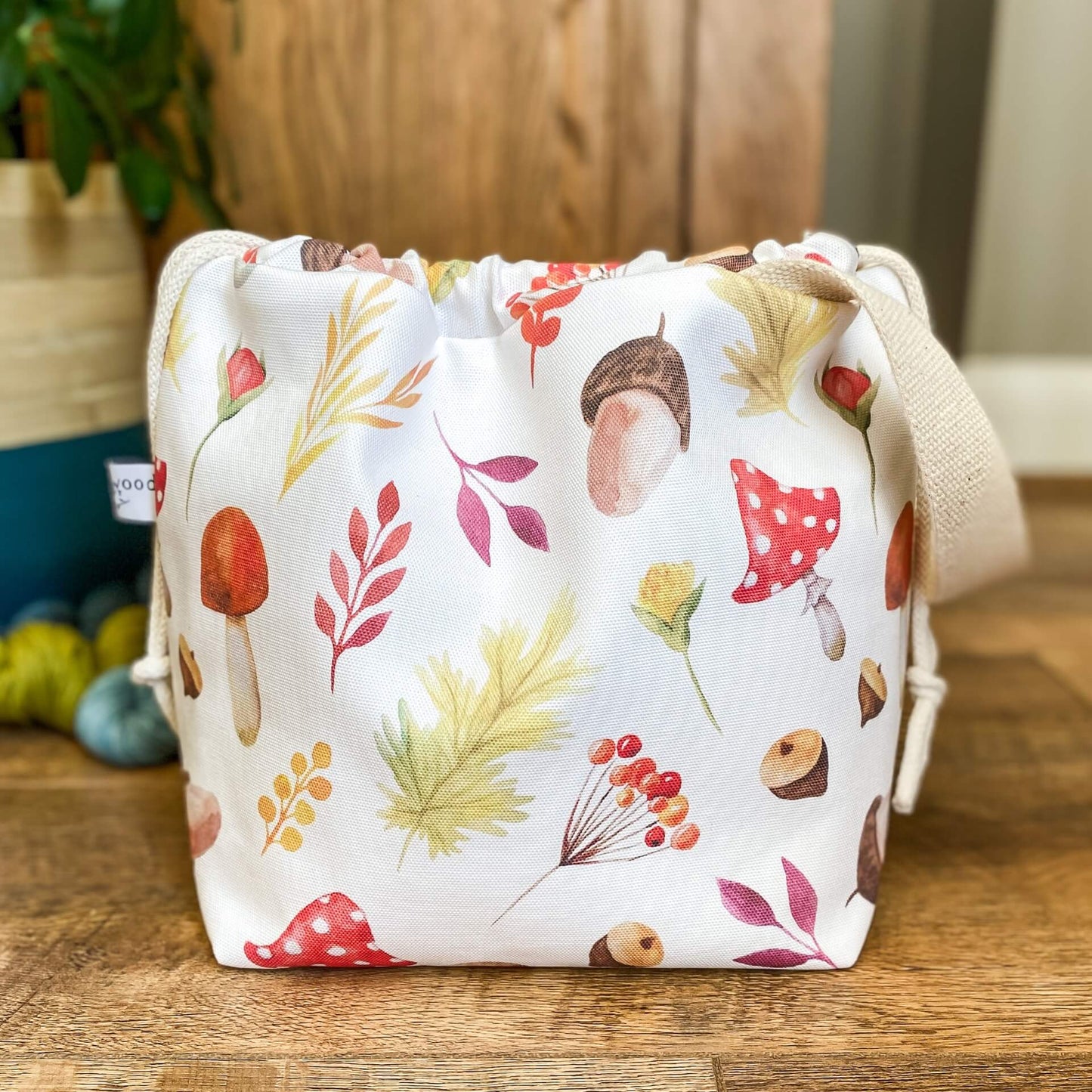 A knitting project bag made using an autumnal print with toadstools and acorns is on the floor next to three skeins of yarn and a houseplant. The bag is pulled closed hiding the contents.