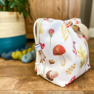A drawstring project bag sits side on showing an Eldenwood Craft label. We also see acorns and toadstools and leaves printed on the bag. The bag sits next to three skeins of yarn and a large houseplant. 