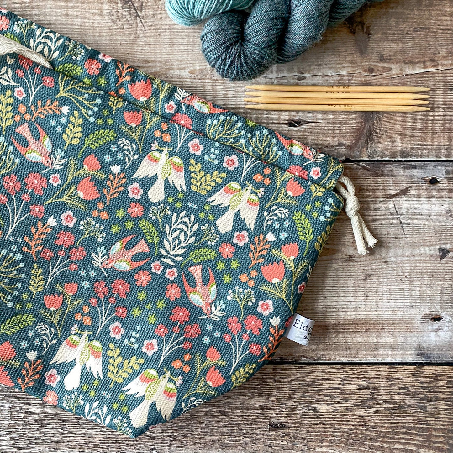 A medium-sized knitting project bag with a bright floral pattern, featuring red and orange flowers and birds on a dark green background. The bag is closed with a drawstring and has fabric handles. Next to it are knitting needles and skeins of yarn on a wooden surface.