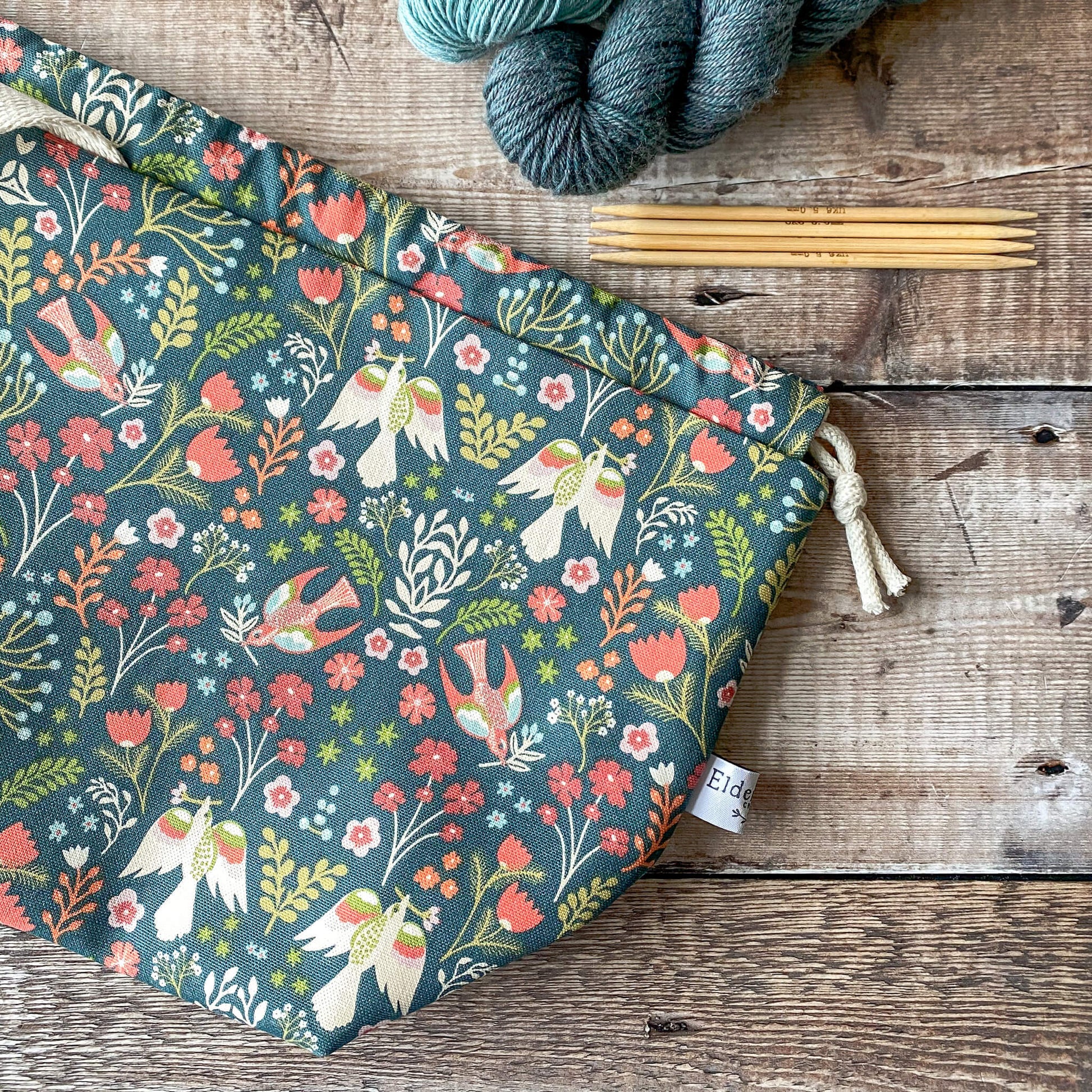 A medium-sized knitting project bag with a bright floral pattern, featuring red and orange flowers and birds on a dark green background. The bag is closed with a drawstring and has fabric handles. Next to it are knitting needles and skeins of yarn on a wooden surface.